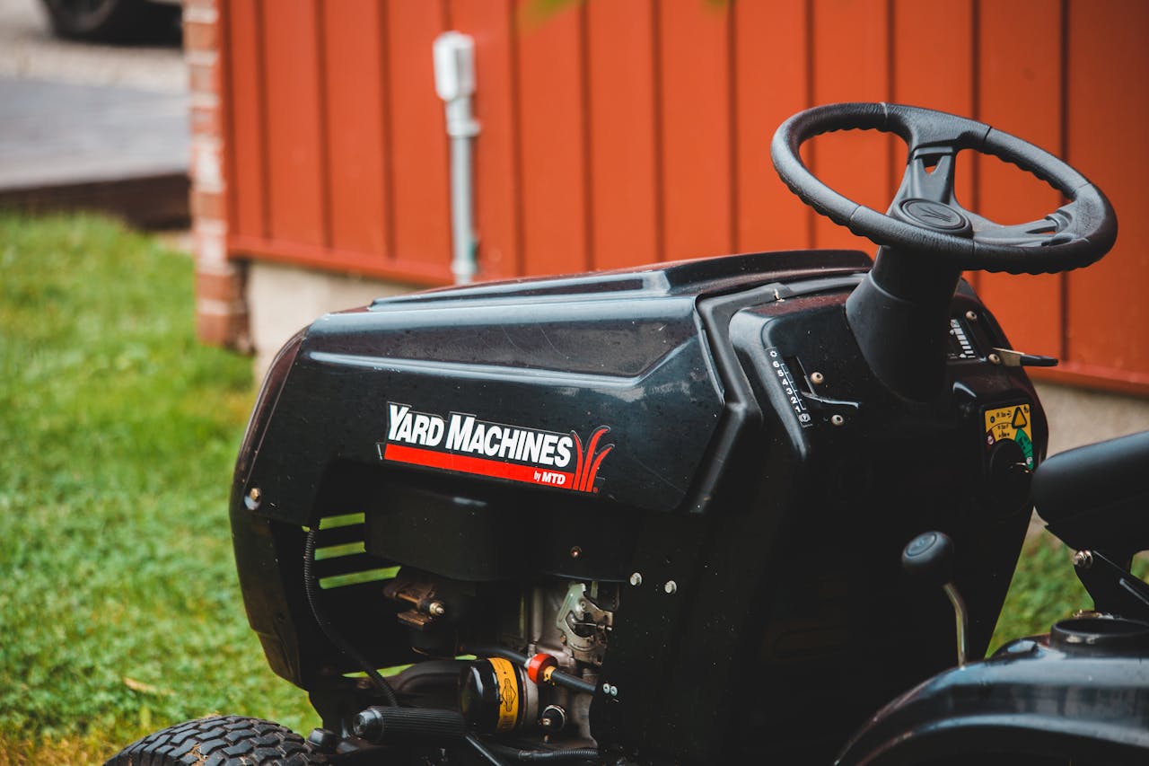 menu-03 Detailed view of a Yard Machines riding lawn mower parked on grassy lawn beside a red shed.